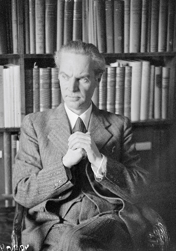 A black-and-white image of an older man in a tweed suit, sitting in a chair in front of a bookcase. He emanates and academic air.