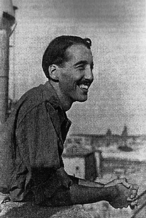 A black-and-white image of a thin main in his thirties, with combed-back hair and a broad smile. He is leaning on a railing on a rooftop, looking out across a city or town.